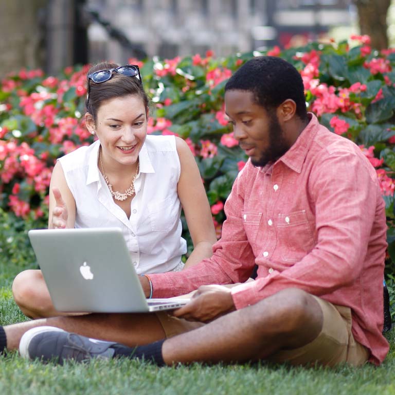Students discuss and look at a laptop while sitting in the grass on campus. 