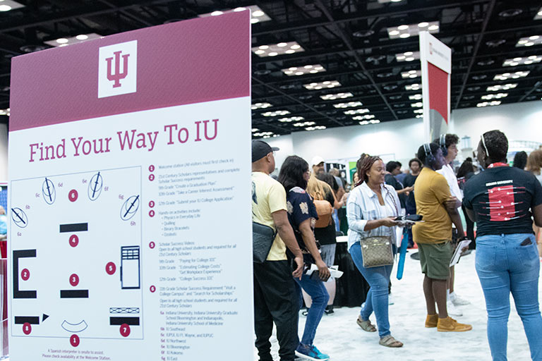An informational board features the IU logo, a title “Find Your Way To IU,” and various icons providing directional guidance. The setting is casual, with diverse attendees. Other similar boards and people can be seen in the background.